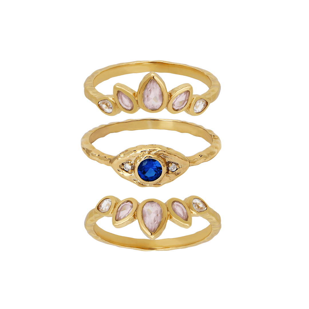 Three gold rings stacked vertically on a white background. The top ring has five pale pink marquise-cut stones, the middle ring has a central round blue stone with small clear stones, and the bottom ring has a design similar to the top ring with pale pink stones.