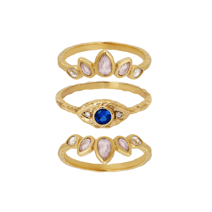 Three gold rings stacked vertically on a white background. The top ring has five pale pink marquise-cut stones, the middle ring has a central round blue stone with small clear stones, and the bottom ring has a design similar to the top ring with pale pink stones.
