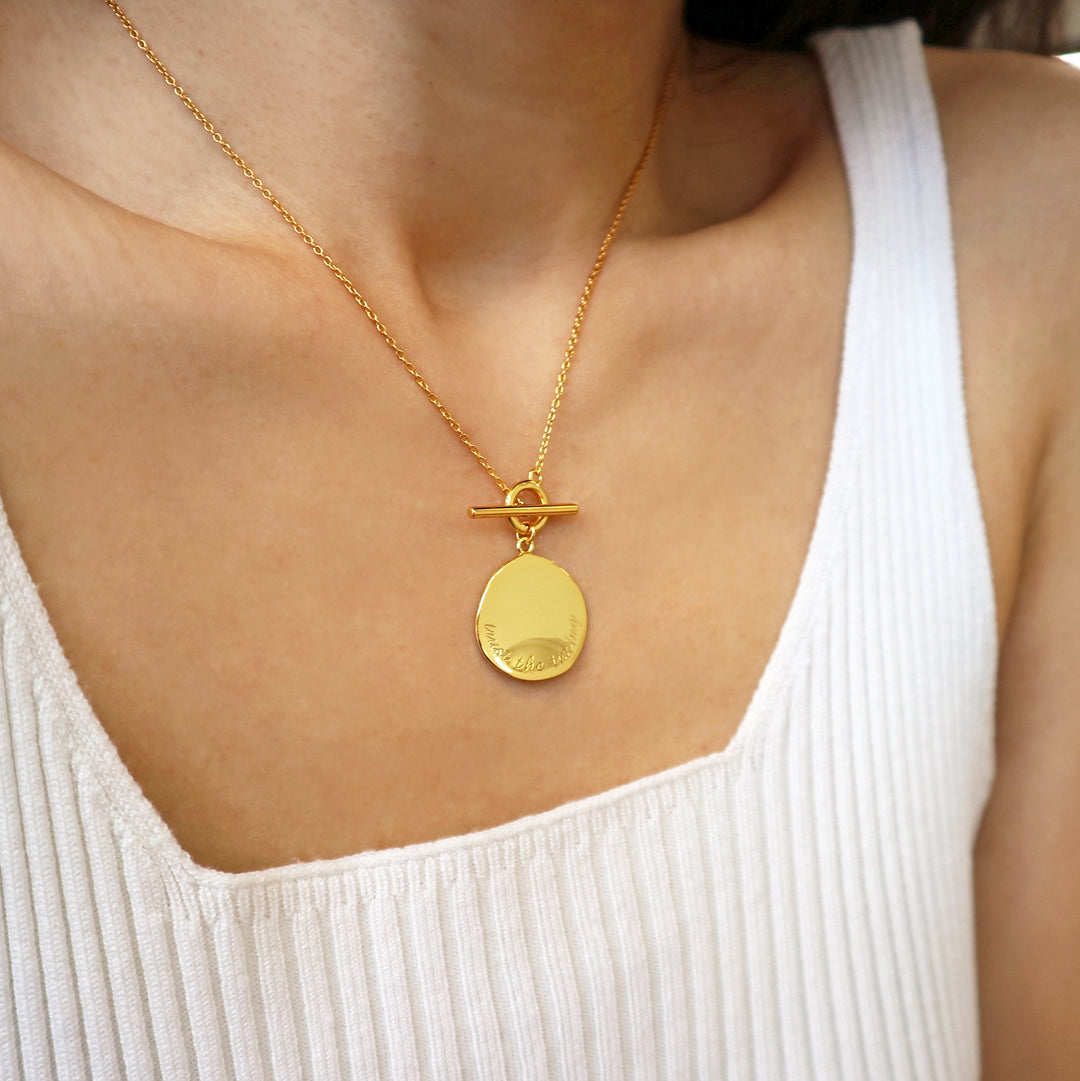 Gold coin necklace with a toggle clasp, engraved with "trust the timing."