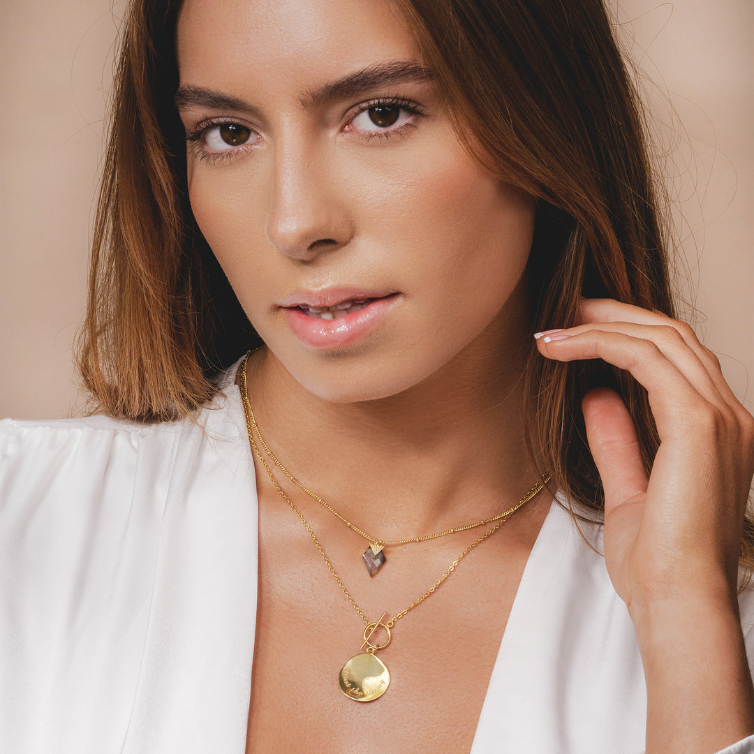 Close-up of a gold coin necklace with "trust the timing" inscription and a functional toggle clasp.