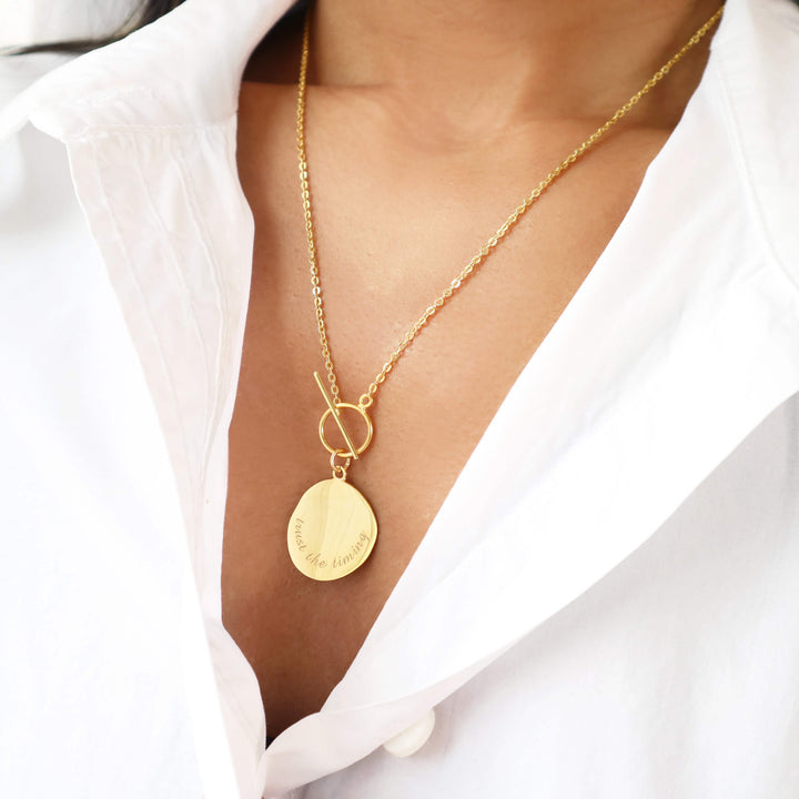 Elegant gold necklace with an engraved coin pendant that reads "trust the timing" and a clasp closure.